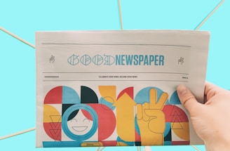 A hand is holding a newspaper with the headline 'Good Newspaper' in a modern design. The newspaper features colorful, abstract artwork with geometric shapes and a smiling face. The background is a solid light blue, creating a clean and minimalistic look.