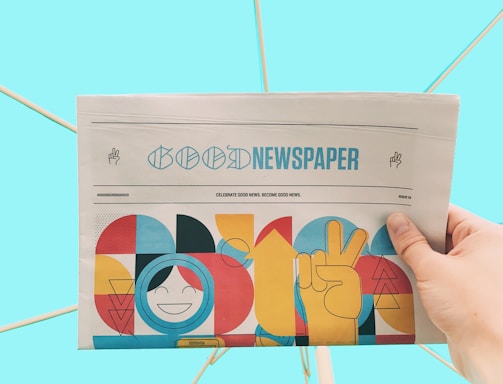 A hand is holding a newspaper with the headline 'Good Newspaper' in a modern design. The newspaper features colorful, abstract artwork with geometric shapes and a smiling face. The background is a solid light blue, creating a clean and minimalistic look.
