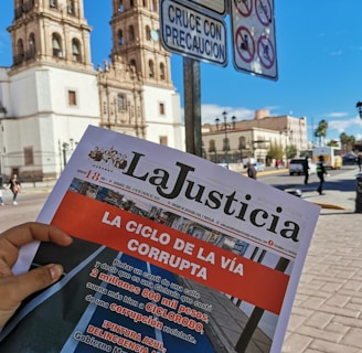 A person's hand holds a newspaper titled 'La Justicia' in front of a historic building with two towers. The newspaper features an article criticizing local government spending on infrastructure. Road signs prohibiting motorbikes, bicycles, and right turns are visible in the foreground, against a bright blue sky.