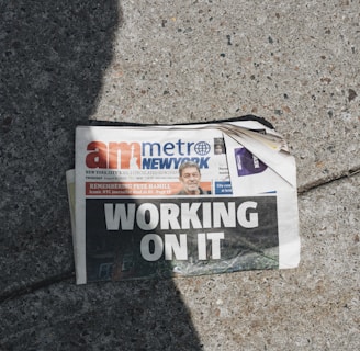 A newspaper lies on a concrete sidewalk with a headline reading 'WORKING ON IT'. The newspaper appears partially crumpled with its pages curling at the corner. It features a mix of photographs and text, including an image of a person in the top section. Sunlight casts a shadow across part of the newspaper and the sidewalk, adding to the scene's contrast.