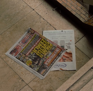 A newspaper with bold headlines and colorful images is placed on a light-colored marble floor. The newspaper features advertisements and various news stories. Adjacent to it is a magazine with an advertisement displayed, partially covered by the newspaper. The surrounding area appears to be part of an architecture with tiles and a darker shadowy corner.